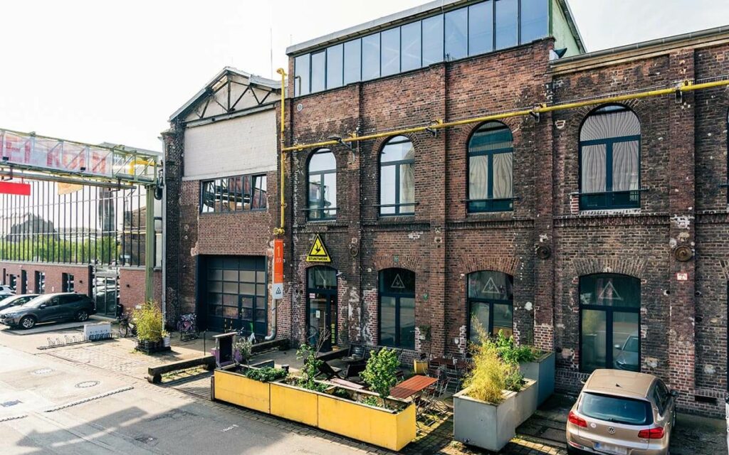 Außenfassade des Stuntwerk Köln Mülheim - altes Ziegelgebäude mit Parkplätzen und Biergarten davor