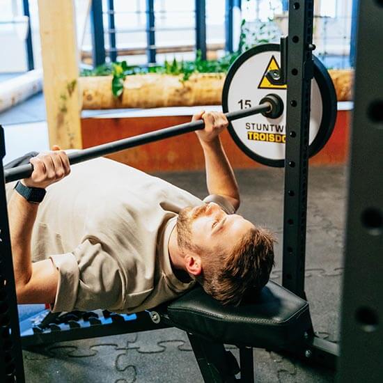 Sportler liegt auf einer schwarzen Trainingsbank und führt Bankdrücken mit einer Langhantel durch. Helle Wände, natürliche Holzelemente und Stuntwerk-Gewichte mit gelbem Logo