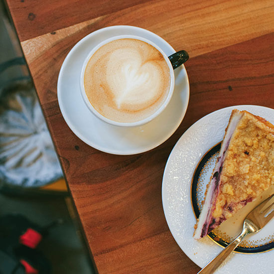 Holztisch von oben - darauf steht eine Tasse Cappuchino und ein Teller mit Streuselkuchen. Unten auf dem Boden liegt ein Chalkbag und Boulderschuhe.