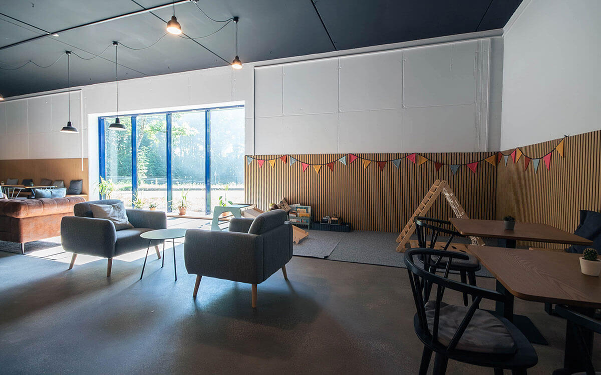 Gemütliches Bistro mit grauen Sesseln, braunem Ledersofa, Holztischen und bunter Wimpelgirlande vor großen Fenstern im Stuntwerk Trostberg.