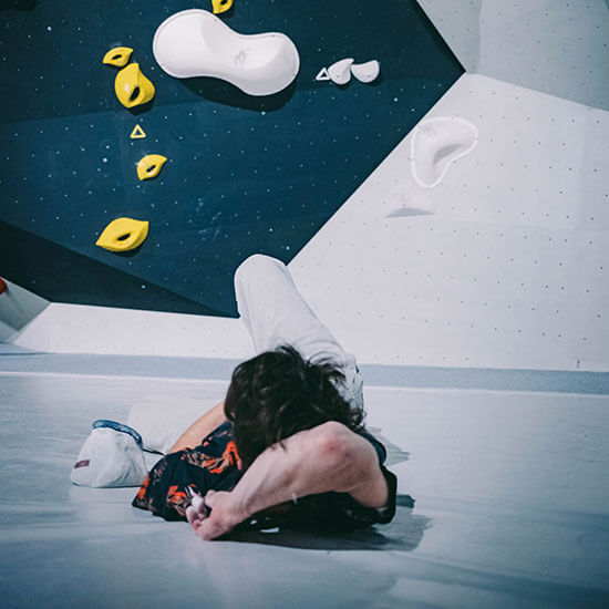 Sportler liegt entspannt auf grauer Matte unter einer schwarz-weißen Boulderwand mit gelben Klettergriffen im Stuntwerk. Dynamische Indoor-Szene nach einem Boulder-Versuch.