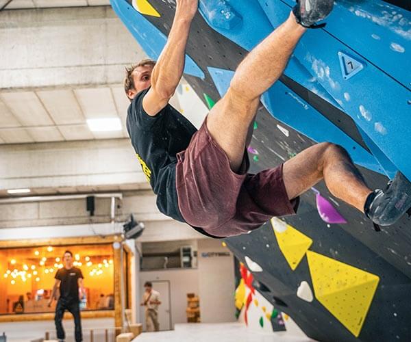 Bouldernder Sportler hängt dynamisch an großen blauen Griffen eines Überhangs in einer hellen Boulderhalle; im Hintergrund sieht man Thekenbereich mit warmen Lichtern und weitere Besucher:innen.