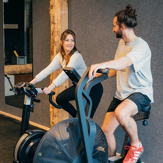 Frau und Mann trainieren nebeneinander auf schwarzen Spinning-Bikes. Warme Beleuchtung, Holzsäulen und graue Wandverkleidung schaffen eine freundliche Trainingsatmosphäre im Stuntwerk.