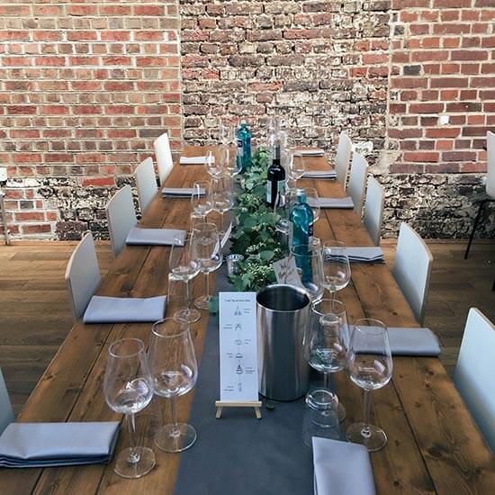 Festlich gedeckter Holztisch mit Weinflaschen, Gläsern und grüner Tischdekoration vor einer rustikalen Backsteinwand – perfekt für Feiern oder Firmenevents.