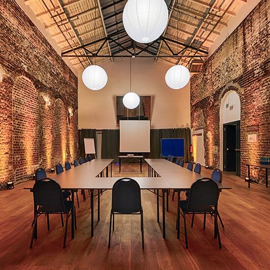 Modern eingerichteter Konferenzraum mit U-förmiger Tischanordnung, Leinwand und stimmungsvoller Beleuchtung vor historischen Backsteinwänden.