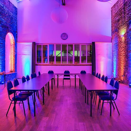 U-förmig aufgestellte Konferenztische in einer beleuchteten Eventhalle mit Backsteinwänden – farbige Lichtakzente in Blau und Lila schaffen eine besondere Atmosphäre.