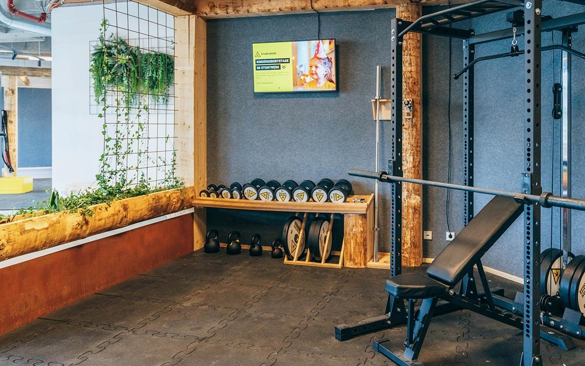 Moderner Fitnessbereich im Stuntwerk Troisdorf mit Holz- und Pflanzenelementen, Hanteln, Rack und Trainingsbank. Warme Atmosphäre mit natürlichen Materialien.