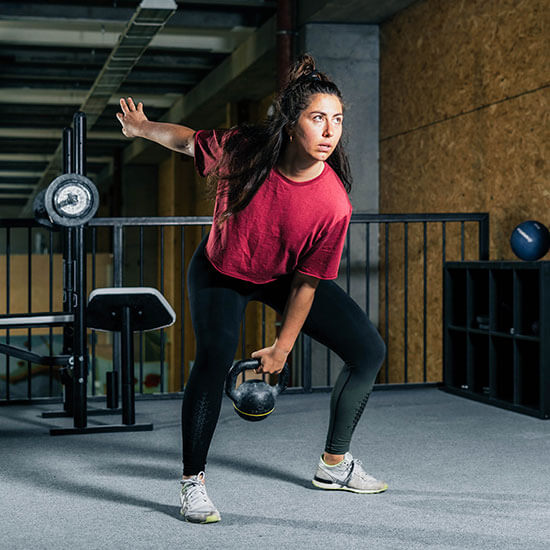 Frau in rotem Oversize-Shirt und schwarzen Leggings schwingt eine schwarze Kettlebell in einem modernen Trainingsraum mit Betonwänden und Metallgeländer im Stuntwerk.