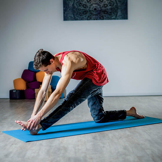 Mann im roten Tanktop dehnt sich auf einer blauen Yogamatte in hellem Raum mit Holzboden und bunten Yogakissen im Hintergrund. Stretching und Mobilitätstraining im Stuntwerk.