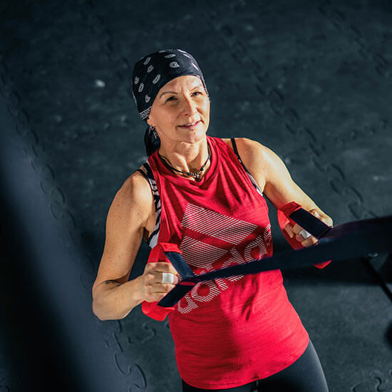 Frau mit schwarzem Bandana und rotem Sporttop trainiert mit TRX-Bändern auf dunklem Boden im Stuntwerk. Konzentration und Körperspannung im Fokus.