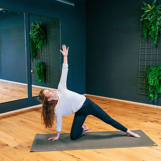 Frau übt im modernen Yoga-Raum des Stuntwerks die Tor-Pose. Holzfußboden, dunkle Wände und vertikale Pflanzen schaffen eine entspannte Studio-Atmosphäre.