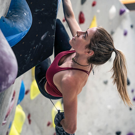Frau in rotem Sporttop klettert fokussiert an großer schwarzen Struktur mit blauen und gelben Klettergriffen im Stuntwerk. Detailaufnahme mit Betonung von Grifftechnik und Körperspannung