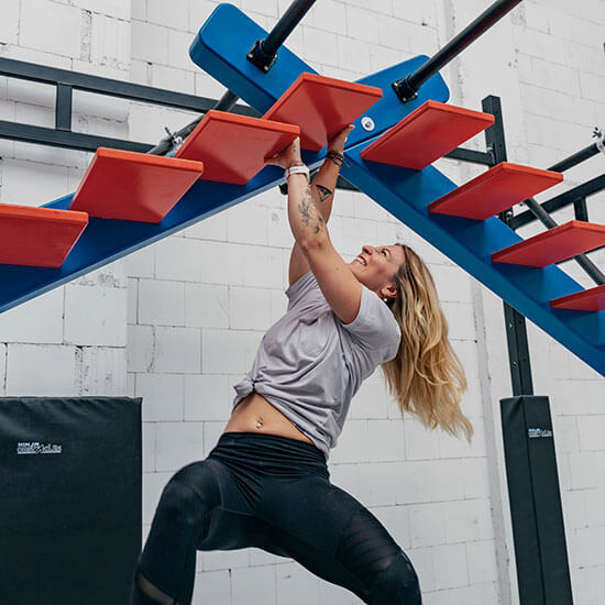 Frau klettert an roter Hindernisleiter im Ninja-Parcours, blaue Trägerstruktur und weiße Backsteinwand im Hintergrund