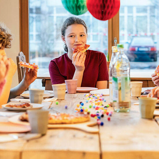 Kinder sitzen an einem festlich geschmückten Tisch mit Pizza und Getränken im Stuntwerk-Bistro. Eine fröhliche Runde genießt gemeinsam den Kindergeburtstag mit bunten Deko-Elementen