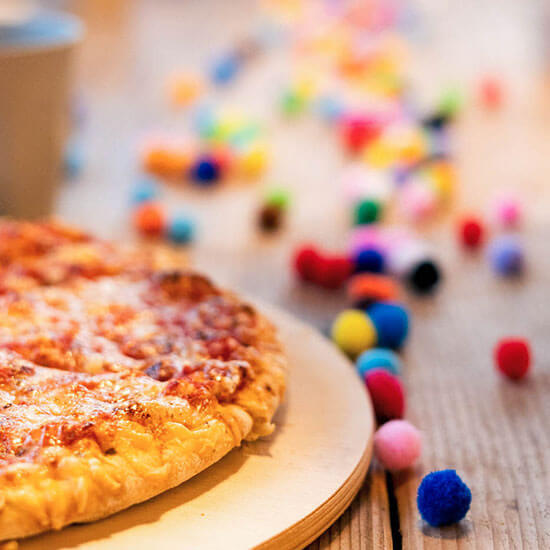 Frisch gebackene Pizza liegt auf einem Holzteller, im Vordergrund bunte Pompons – festliche Stimmung bei einem Kindergeburtstag im Stuntwerk.