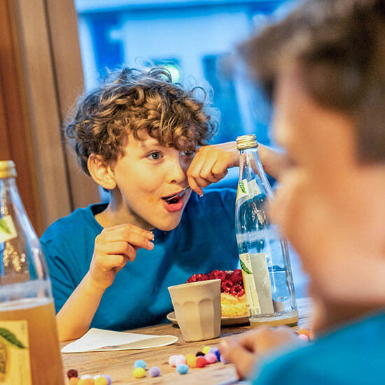 Junge mit lockigen Haaren sitzt lachend am Tisch im Stuntwerk und genießt ein Stück Himbeerkuchen. Neben ihm stehen Glasflaschen mit Wasser und bunte Pompons liegen verstreut auf dem Holztisch – entspannte Pause nach dem Bouldern.