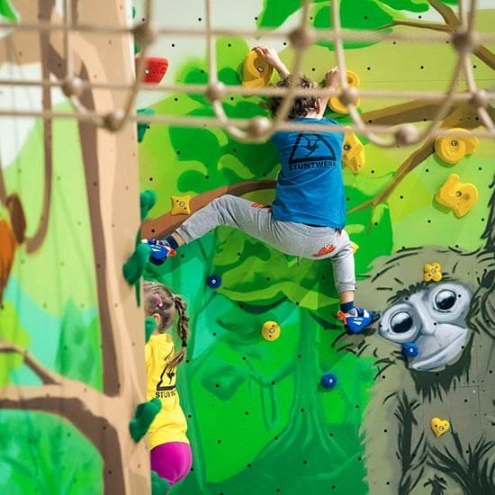Zwei Kinder klettern an einer farbenfrohen Dschungel-Kletterwand im Stuntwerk, bemalt mit grünen Blättern und einem gezeichneten Affen. Beide tragen Stuntwerk-Shirts und nutzen bunte Klettergriffe – spielerisches Bouldern mit Fantasie und Abenteuer.