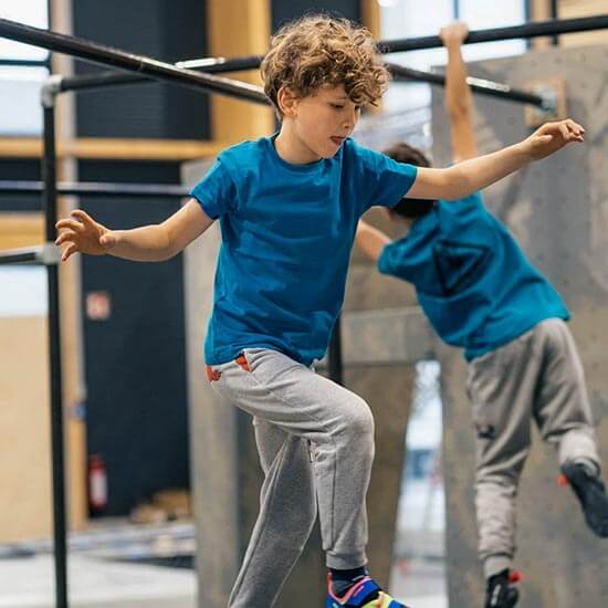 Junge balanciert konzentriert auf einer Parkour-Stange im Stuntwerk. Im Hintergrund trainieren andere Kinder an den Hindernissen. Der Junge trägt ein blaues T-Shirt und graue Jogginghose – voller Fokus im Kids-Training