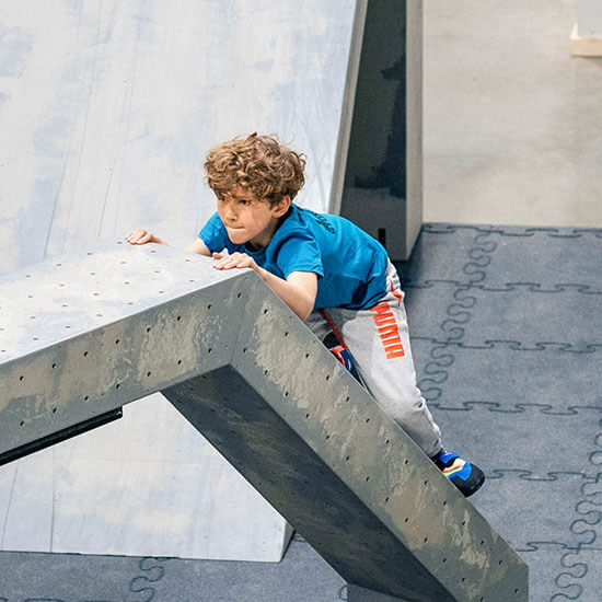 ind klettert konzentriert auf einem geneigten Hindernis im Parkourbereich des Stuntwerks. Mit sportlicher Körperspannung und Fokus erklimmt es das Hindernis – Spaß und Herausforderung im Kindertraining.