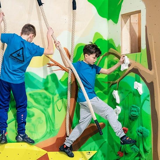 „Zwei Jungen schwingen sich an dicken Seilen über eine Kletterwand im Stuntwerk. Die Wände sind bunt bemalt mit Bäumen und Tieren – spielerische Mischung aus Bouldern und Ninja-Training für Kinder.