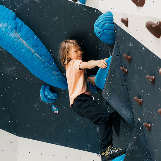 Kind mit orangefarbenem T-Shirt und schwarzen Kletterhosen bouldert an einer schwarz-weißen Wand mit großen blauen Griffen im Stuntwerk. Konzentration und Spaß im Mixed-Boulderbereich.