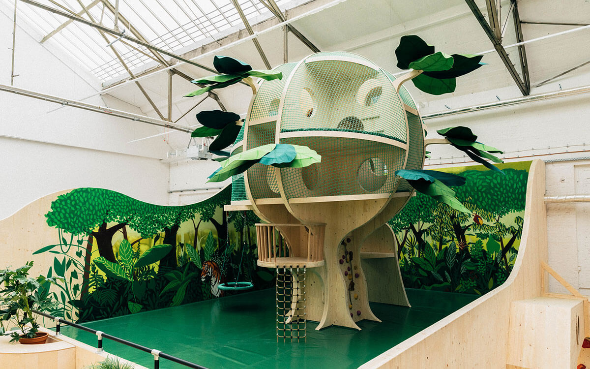 Großer Kletterbaum im Kinderbereich des Stuntwerks Köln Mülheim. Die kugelförmige Holzkonstruktion mit Netzen und grünen Blättern erinnert an ein Baumhaus und bietet mehrere Ebenen zum Spielen, Klettern und Entdecken – mitten im Dschungel-Design.