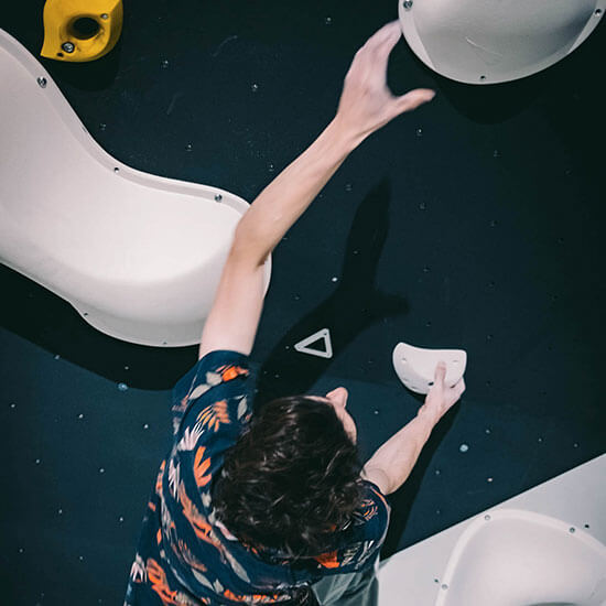 Kletterer in bunter, dunkelblauer Shirt mit orangenem Muster greift an einer schwarzen Boulderwand nach weißen Klettergriffen. Überkopf-Perspektive mit Fokus auf Bewegung und Grifftechnik.