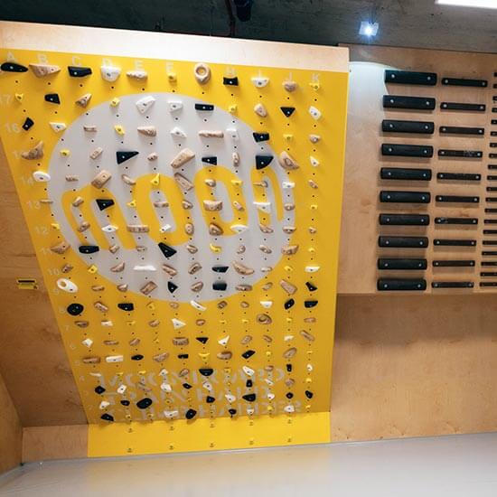 Gelb-weiße Trainingswand mit dem Moonboard-Logo, bestückt mit hellen und schwarzen Klettergriffen. Holzverkleidung und präzise angeordnete Griffstruktur für Technik- und Krafttraining im Stuntwerk.