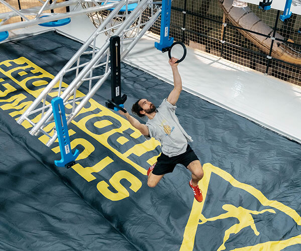 Ein Mann mit Bart, im grauem T-Shirt und mit roten Sportschuhen hangelt sich von Hindernis zu Hindernis. Gerade hält er sich an einem Ring fest. Unter ihm befindet sich ein schwarzer Airbag mit dem gelben Aufdruck "Stuntwerk Troisdorf"