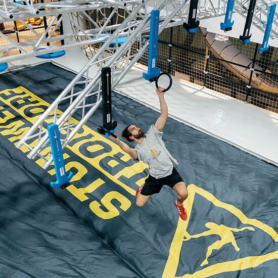 Blick auf den Ninja Frame im Stuntwerk Troisdorf. Ein Mann hangelt sich mit Ringen durch den Parcour. Unter ihm ein Luftkissen für eine weiche Landung.