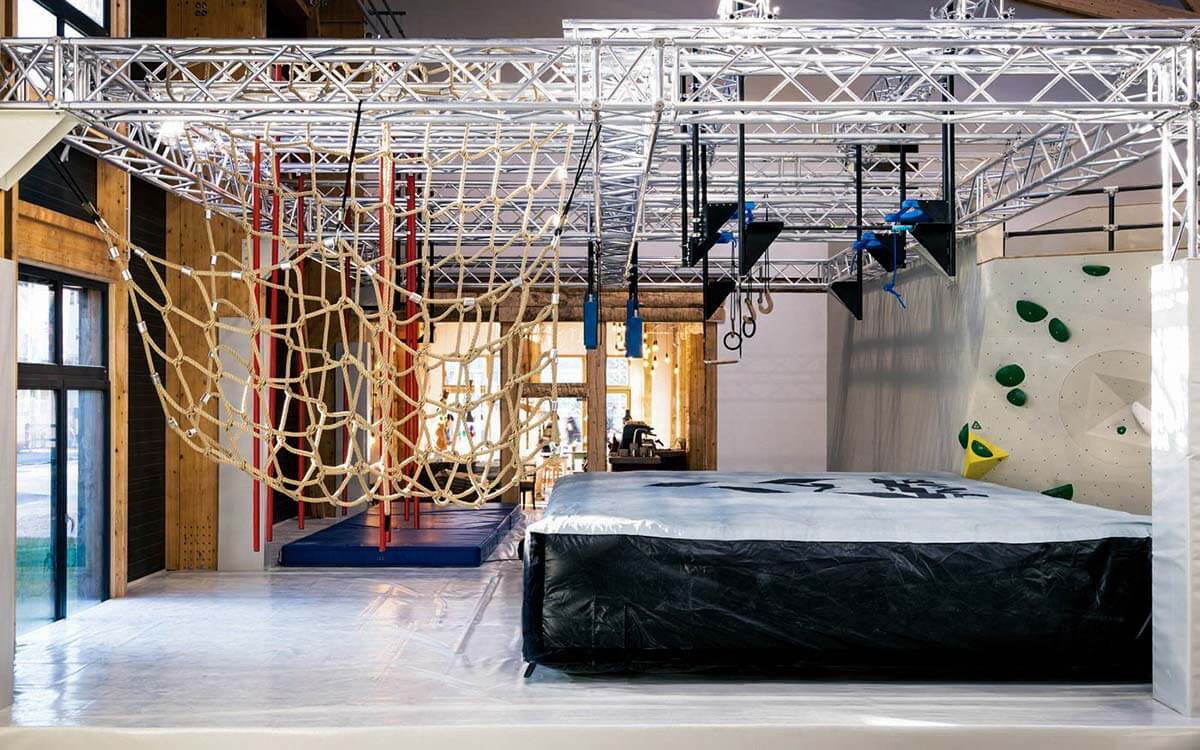 Großer Ninjaframe mit Blick von/ins Bistro. Es sind verschiedene Hangelhindernisse, wie Netz, Stangen und Ringe aufhängt. Eine dicke Fallschutzmatte und ein Luftkissen schützen vor Verletzungen