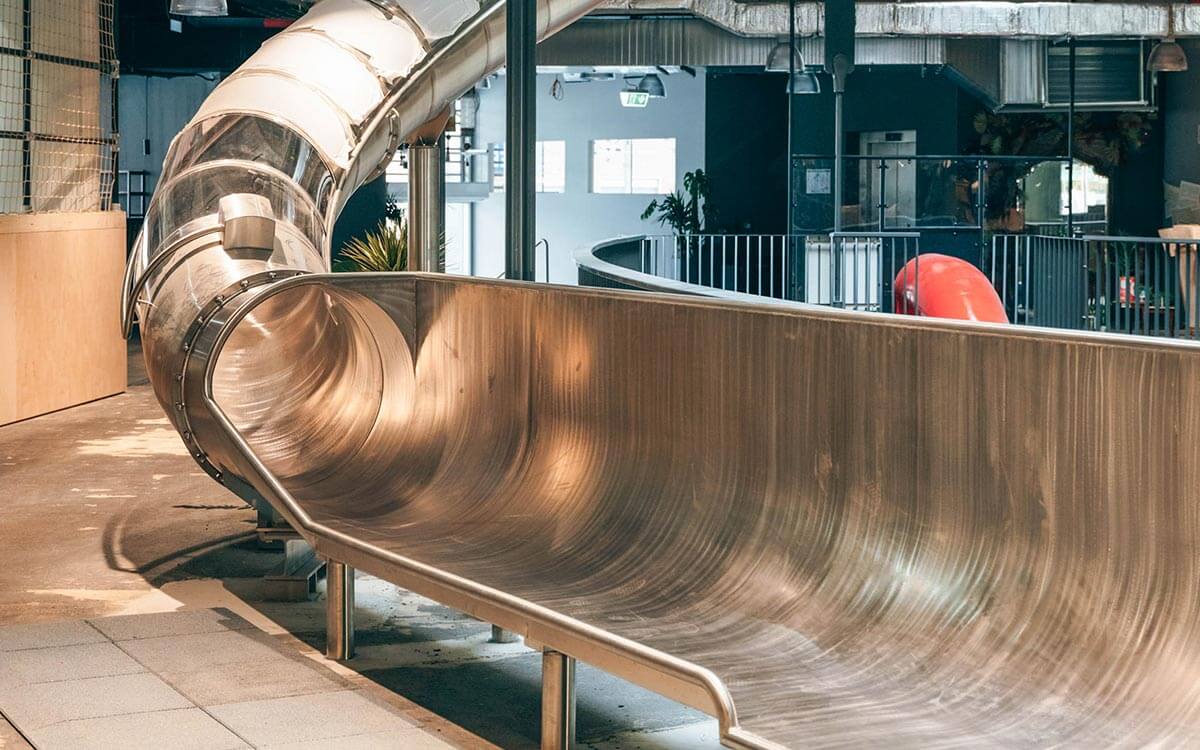 Glänzende Edelstahlrutsche im Stuntwerk Troisdorf mit transparentem Einstiegstunnel. Die moderne Indoor-Rutsche verbindet die oberen Ebenen der Halle und sorgt für Spaß und Action beim Abstieg.
