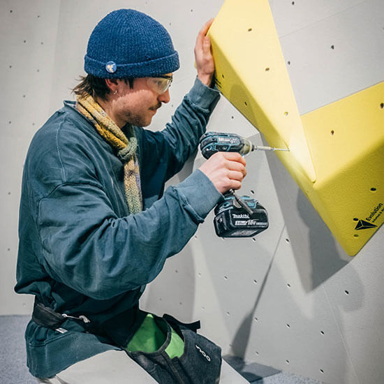Routensetzer befestigt ein gelbes Boulder-Volumen an einer hellgrauen Kletterwand mit Akkuschrauber im Stuntwerk. Er trägt eine blaue Mütze, Schutzbrille und dunklen Pullover