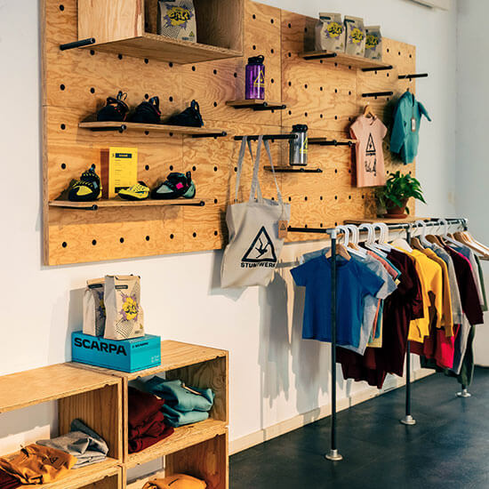 Heller Shopbereich im Stuntwerk mit Holzwänden, Pflanzen und farbenfrohen Shirts. Auf den Regalen stehen Kletterschuhe, Trinkflaschen und Merchandise-Artikel.