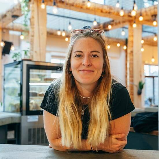 Mitarbeiterin im Stuntwerk an der Theke - sie lächelt freundlich in die Kamera, ist entspannt auf die Theke gelehnt. Ihre blonden Haare fallen locker über ihre Schultern, auf dem Kopf trägt sie eine Sonnenbrille. Im Hintergrund sieht man eine Auslage mit Kuchen, die mit Holzbalken gestaltete Decke wird durch Glühbirnen in ein weiches Licht getaucht.