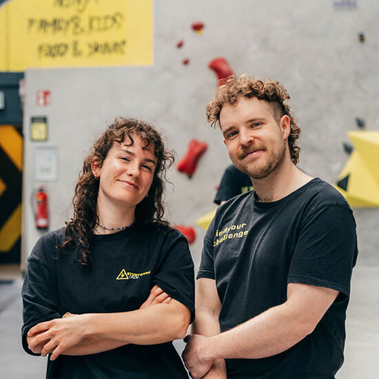 Zwei Mitarbeitende des Stuntwerks stehen lächelnd nebeneinander in der Boulderhalle. Beide tragen schwarze Stuntwerk-Shirts und strahlen Selbstbewusstsein und Teamgeist aus. Im Hintergrund sind Boulderwände mit roten und gelben Griffen zu sehen.