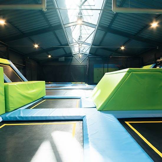 Großer Trampolinpark im Stuntwerk mit mehreren Sprungfeldern, grünen Hinderniselementen und blauen Sicherheitsmatten unter hoher Hallendecke.