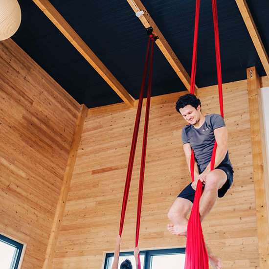 Teilnehmer hängt lachend im roten Vertikaltuch im hellen Holzraum des Stuntwerks. Der Kurs vereint Krafttraining, Körperspannung und Spaß an der Bewegung in der Luft.