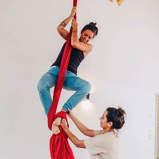 Trainerin unterstützt Teilnehmerin beim Vertikaltuch-Training im Stuntwerk. Die rote Tuchbahn hängt von der Decke, während die Teilnehmerin lachend Balance und Kraft trainiert.